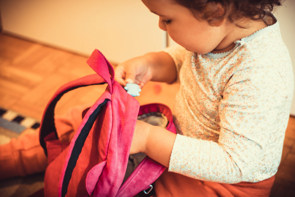Small girl sitting on the floor and packing her backpack while preparing for first day at kindergarten.