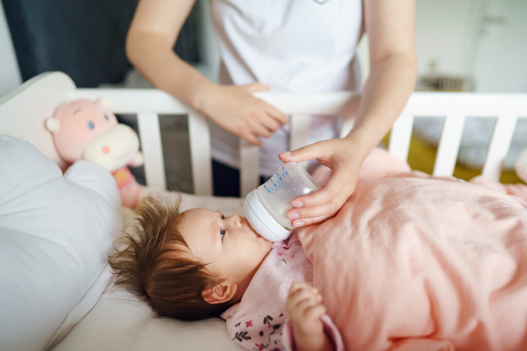 Front view of caucasian woman mother taking care of his newborn baby child two months old bottle feeding with breast or formula milk while child is lying in cradle at home selective focus copy space