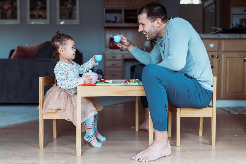 Profile view of a dad and his toddler daughter sitting at a child size table and doing a celebratory toast with toy teacups while playing together at home.