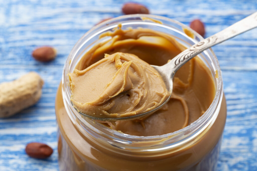 Peanut butter in an open jar and peanuts in the skin are scattered on the blue table. Space for text. Closeup.