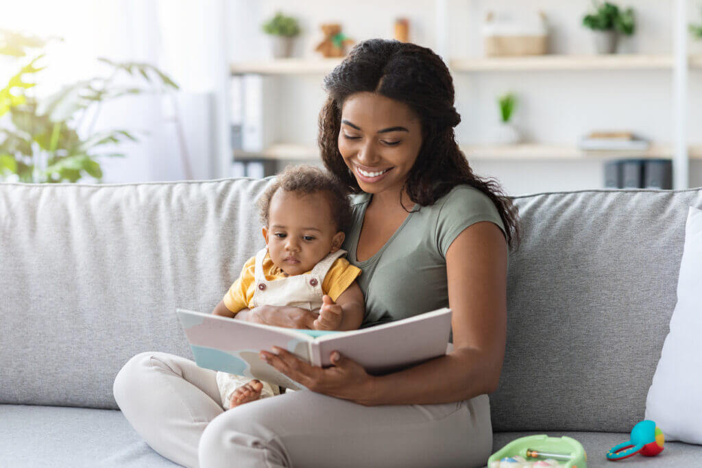 Portrait Of Caring Black Mom Reading Book To Cute Little Baby While They Relaxing On Couch At Home Together, Closeup Shot Of Loving African American Mother Bonding With Her Infant Child, Copy Space