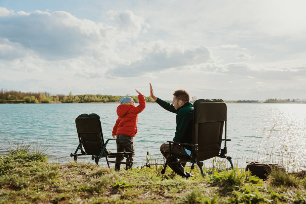 Little boy giving high five to father sitting on camping chair at riverbank