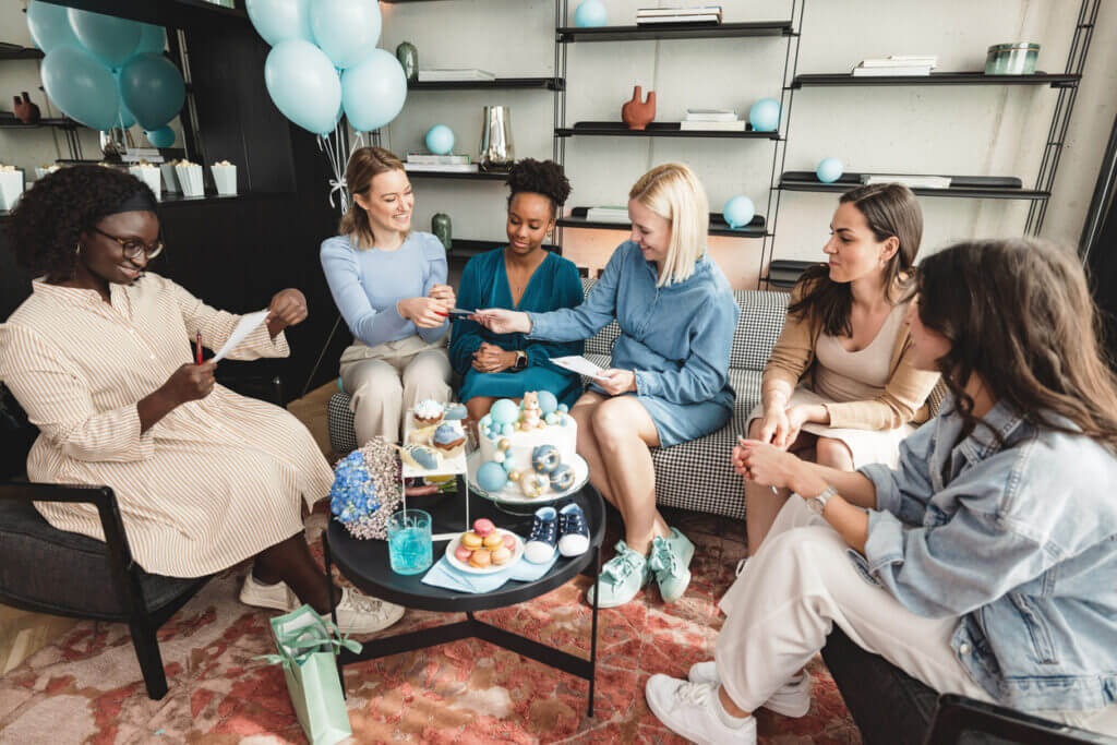 A multiracial group of female friends having fun and playing games at a baby shower celebration. They are about to have a quiz on how much they know the mother-to-be. They are located in a beautifully decorated living room filled with balloons and sweet treats. Fun activities at a baby shower celebration.