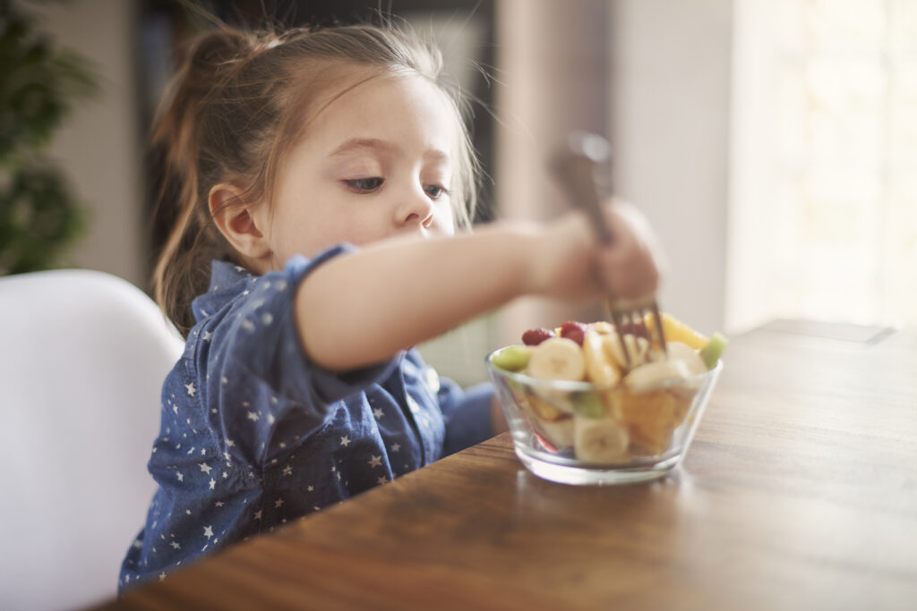 Young girl sitting at a table eating fruit salad with a fork