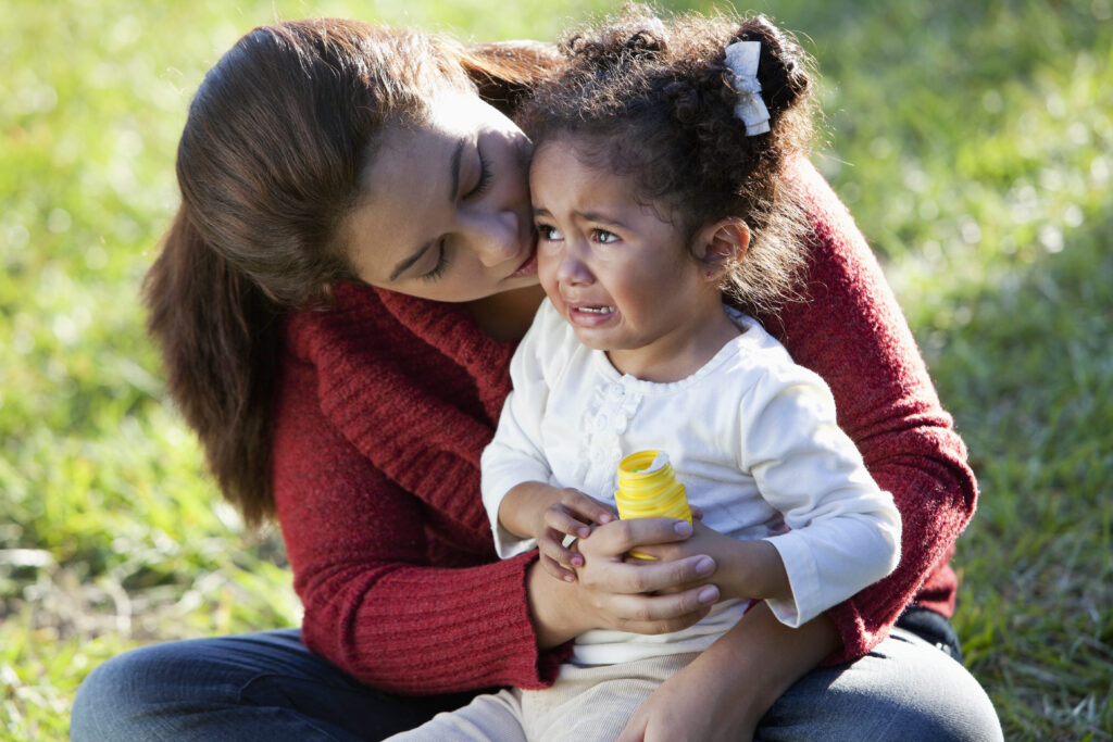 A cute little two year old hispanic girl crying, sitting in her mother's lap. Mom is holding her, trying to comfort her. They are sitting on the grass outdoors.