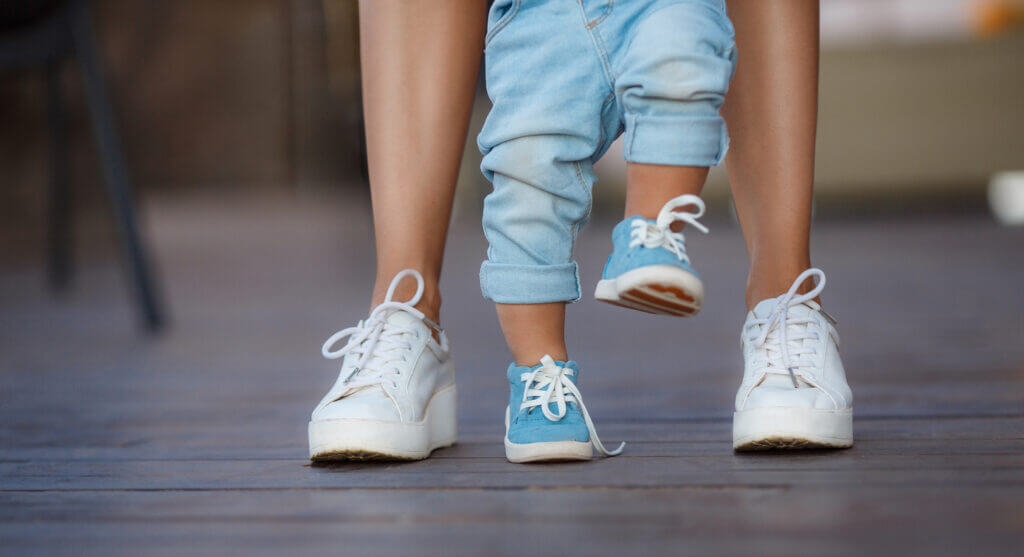 Mom in white sneakers with a young son, dressed in blue pants and blue shoes, a warm summer day and learn to walk in the street, the first steps, the mother maintains her son.