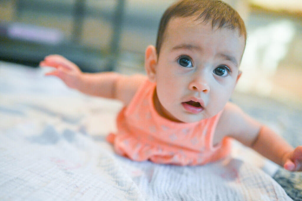 Cute little baby girl around 3 months old practices tummy time for strengthening back and neck muscles. She lays on a baby blanket on the floor on her stomach and reaches a baby milestone. Cute Latina baby child