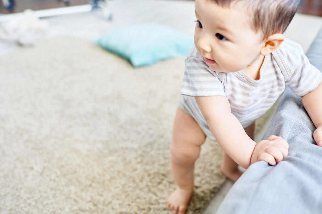 Full-length portrait of adorable baby boy holding on cozy sofa while learning to walk, interior of spacious living room on background